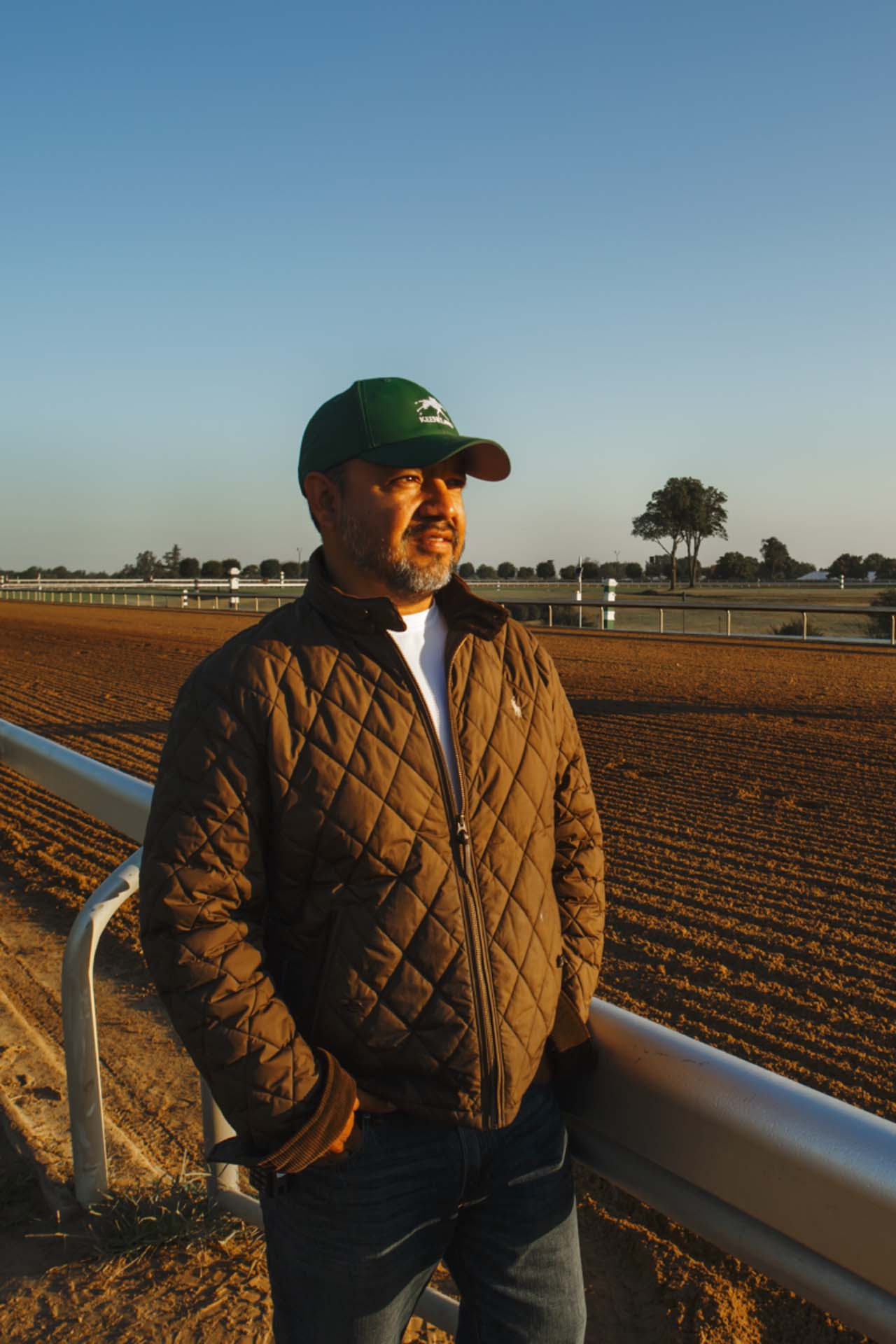 A photo of Alfredo Laureano looking off into the distance while standing by the dirt track at sunset. He is an older Hispanic man with graying chin stubble. He is wearing a green Galloprush-brand hat and a brown Galloprush-brand jacket overtop a white shirt.